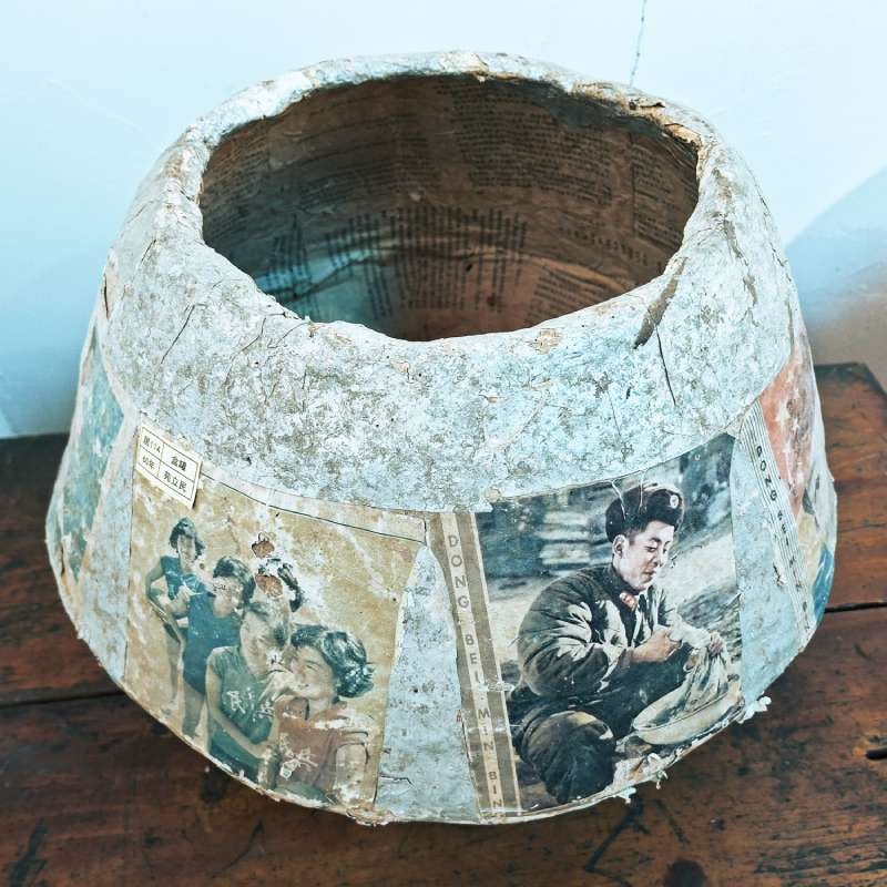 Round, worn concrete planter wrapped in vintage newspaper clippings with a hollow opening at the top.