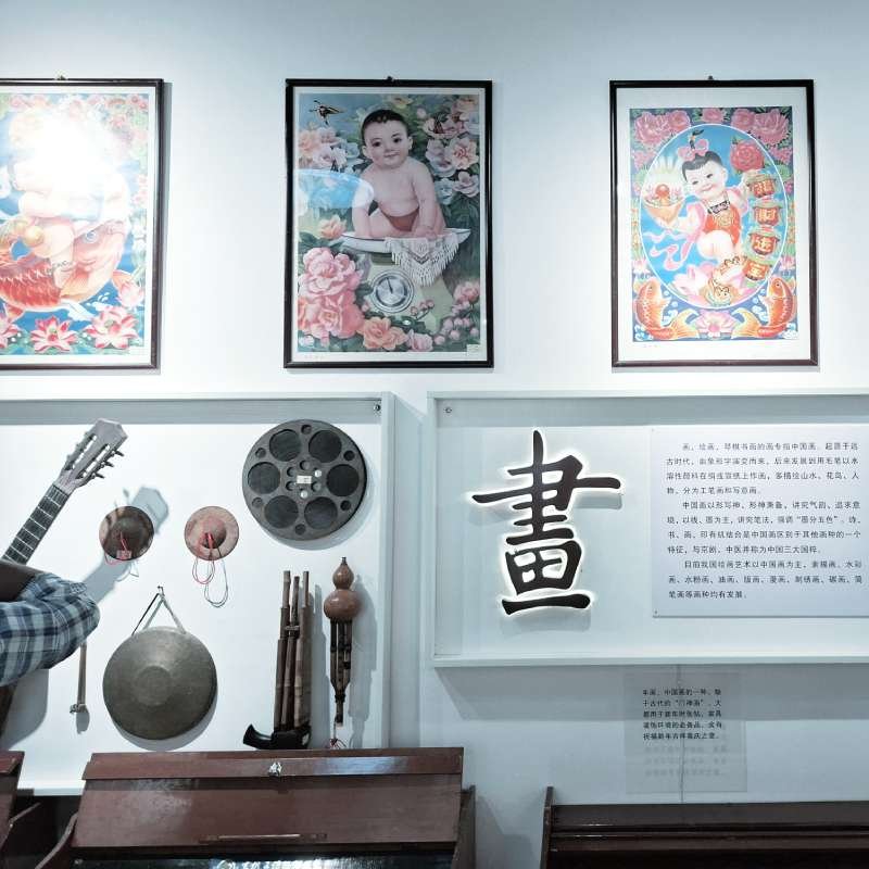 Museum display: three framed colorful baby deity posters above a wall with traditional instruments and a large Chinese character panel.