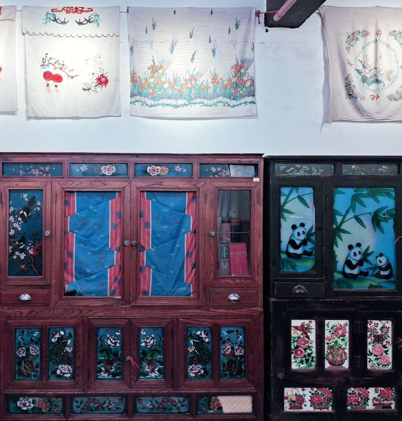 Two vintage wooden display cabinets with glass doors and floral/nature panels, topped by embroidered white fabrics on a wall.