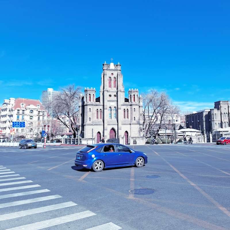 Blue hatchback driving through a spacious city square with a Gothic stone building in the background on a sunny day.