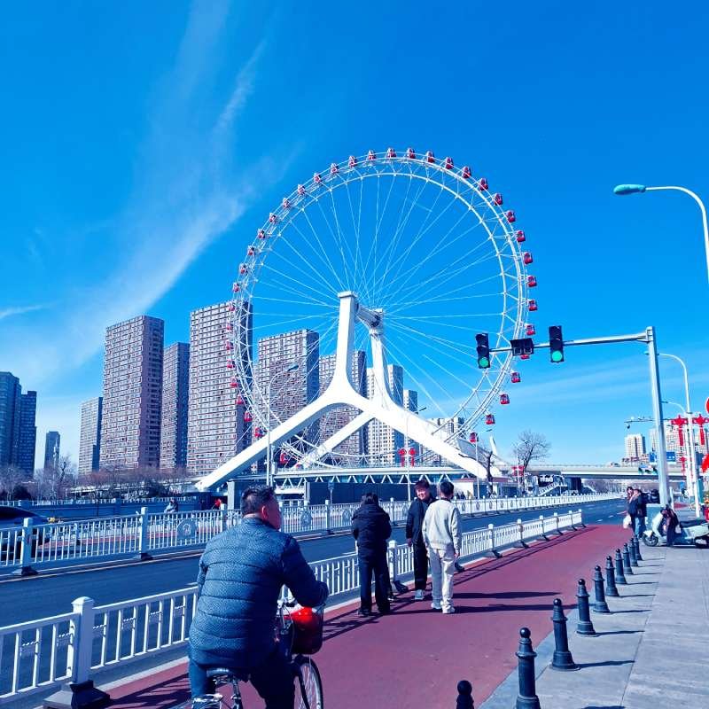 Large white Ferris wheel beside a city skyline, with pedestrians on a riverfront promenade.