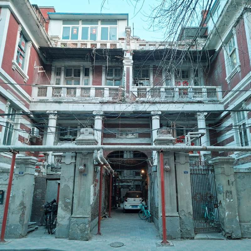 Three-story brick building with decorative white columns and a gated archway; a parked white car and bicycles in the narrow alley beneath the structure