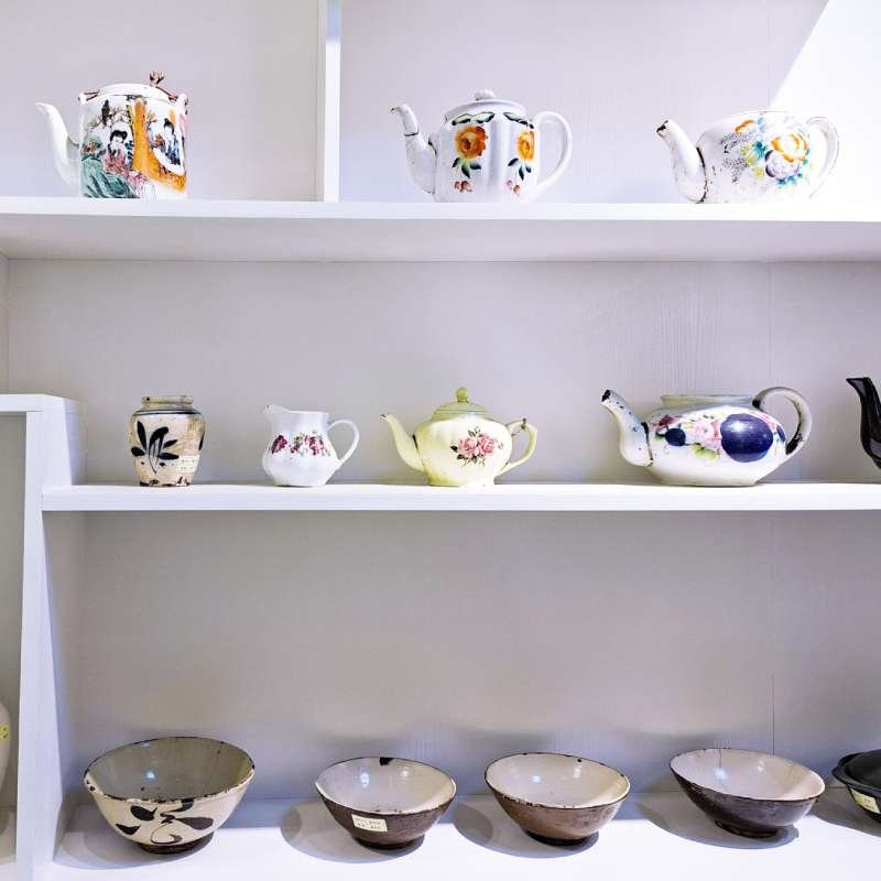 White shelving unit with decorative teapots—floral and scenic patterns—on three shelves and a row of bowls on the bottom shelf.