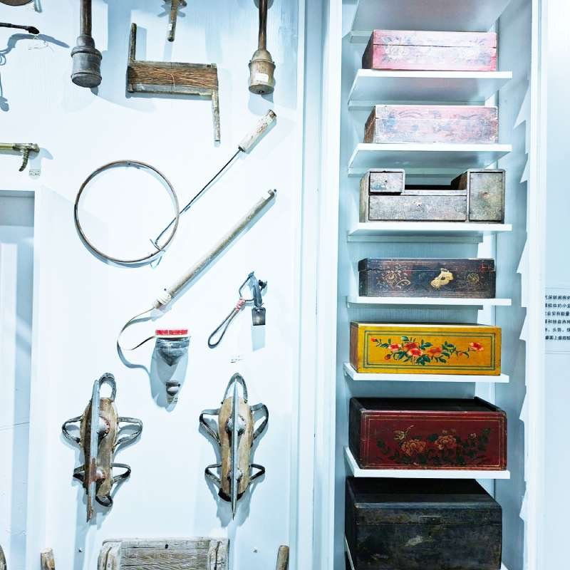 Vintage woodworking tools mounted on a white wall next to shelves filled with painted boxes in a museum display.