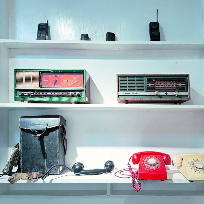 Vintage shelf displaying two old radios and a selection of retro telephones and a black carrying case on the bottom shelf