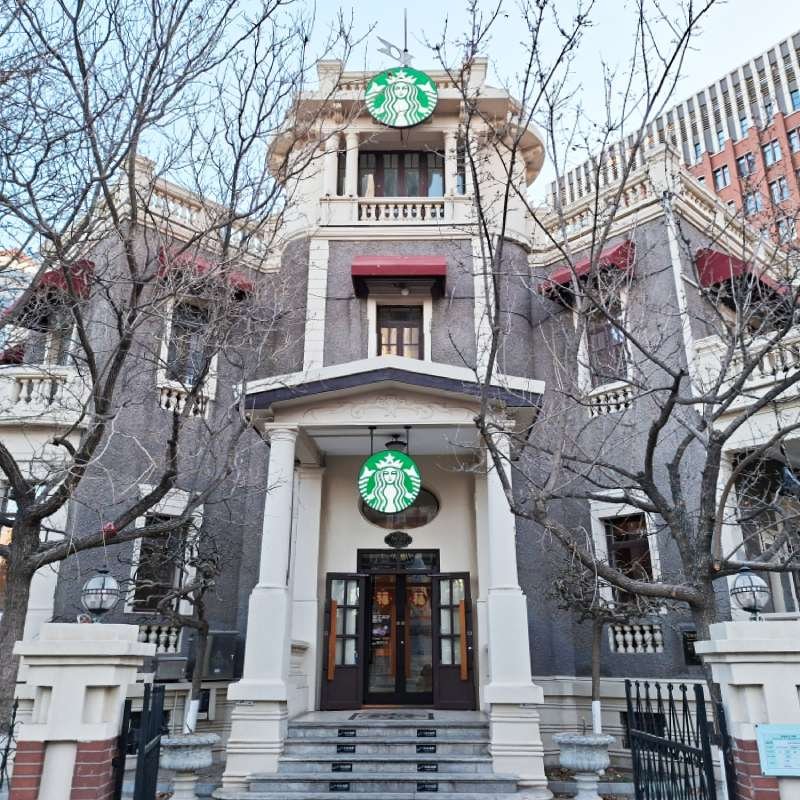 Historic-style building housing a Starbucks cafe, two signs with the green Starbucks logo visible above entrance and on balcony, leafless trees framing the facade.