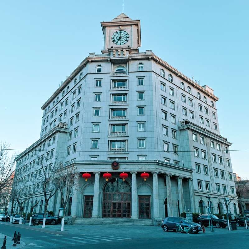 Large grey institutional building with a clock tower, several windows, and red lanterns hanging at the entrance; cars parked along the street.