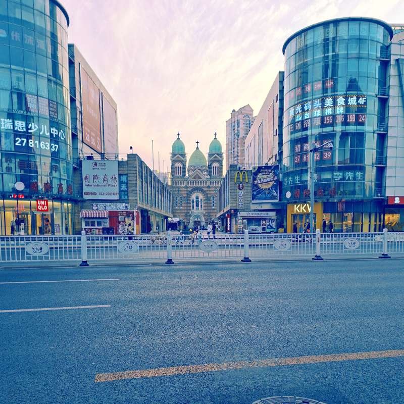 Wide city street lined with glass-fronted shops, leading to a cathedral-like building with two green domes in the distance.