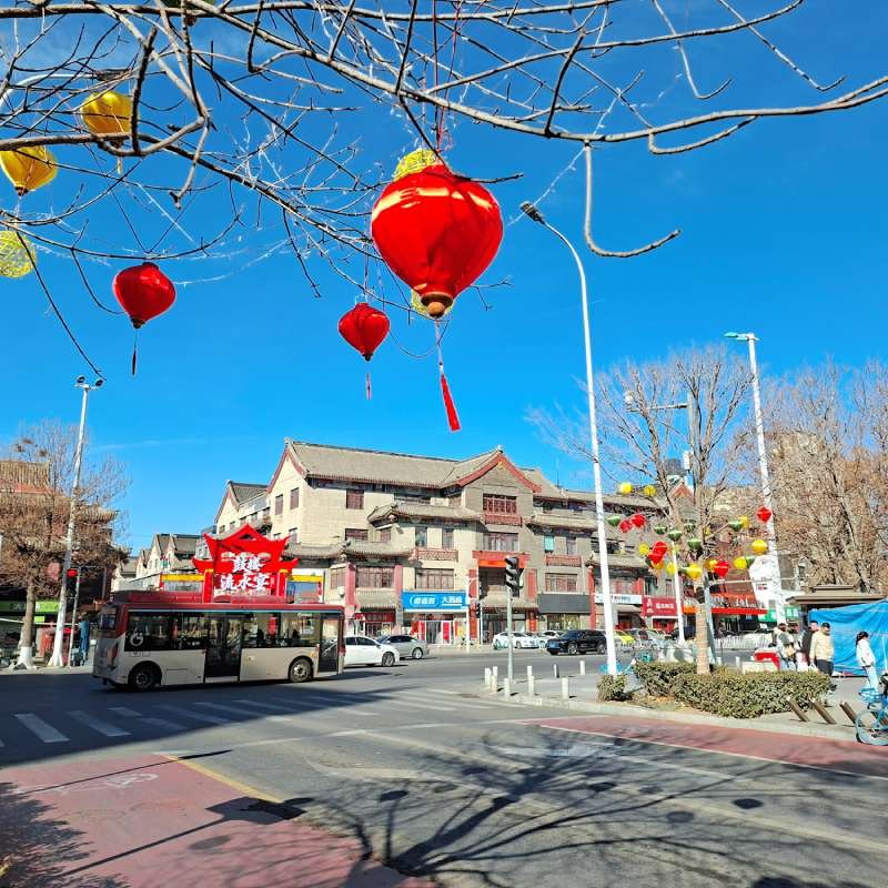 Red lanterns hang from tree branches over a busy street with traditional Chinese buildings and a white bus nearby.