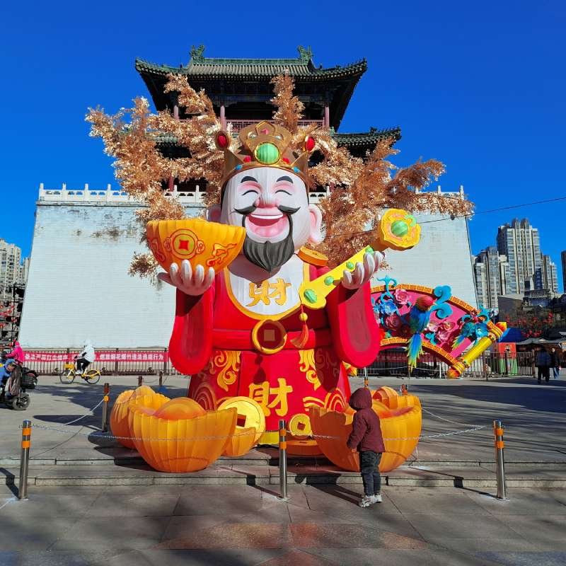 Large colorful statue of the Chinese God of Wealth holding a gold ingot, surrounded by orange pumpkins in a city plaza.