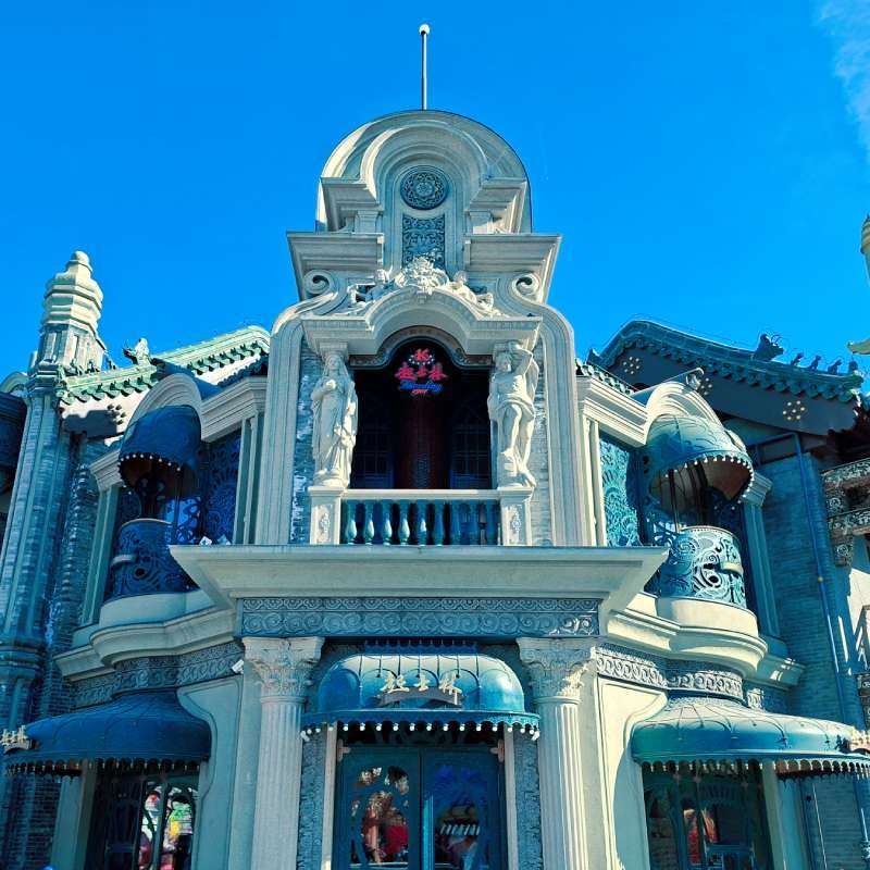 Ornate blue-and-white building with decorative arches, statues, and curved roofs against a bright blue sky (a themed architectural facade). Themed storefront or attraction façade.