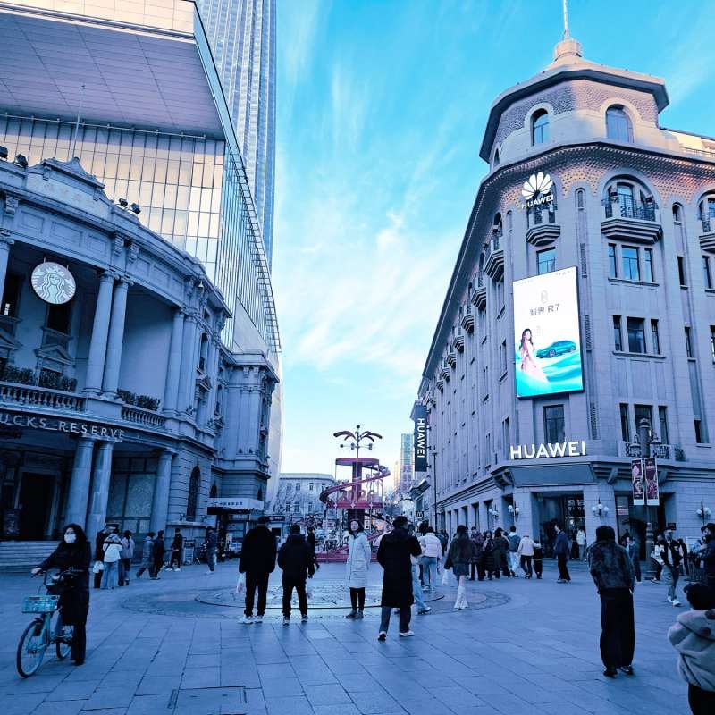 Busy city street with Huawei store on the right and Starbucks Reserve on the left; pedestrians walk under a clear blue sky.