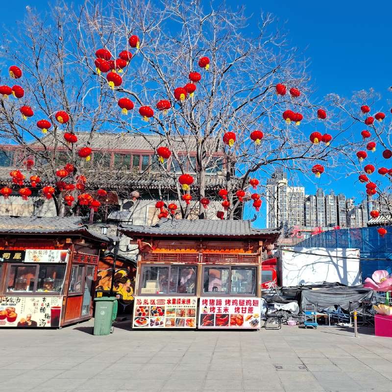 Row of red Chinese lanterns hanging from bare trees over street food stalls in a plaza, blue sky and apartment buildings in the background.