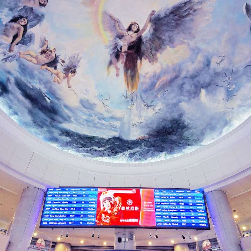 Circular ceiling mural of winged angels above ocean waves, with a large digital departure board below in a transit terminal.