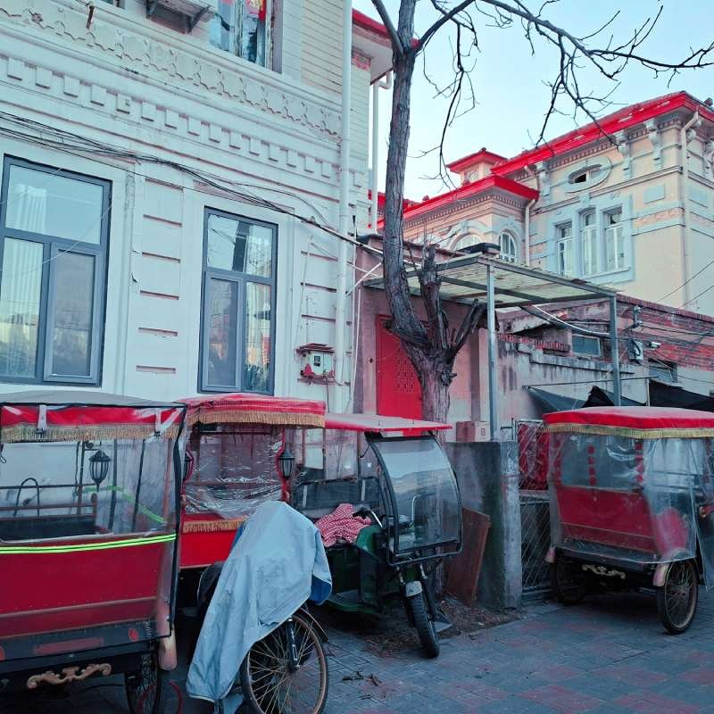 Row of red-covered rickshaws parked along a sidewalk beside white and cream buildings.