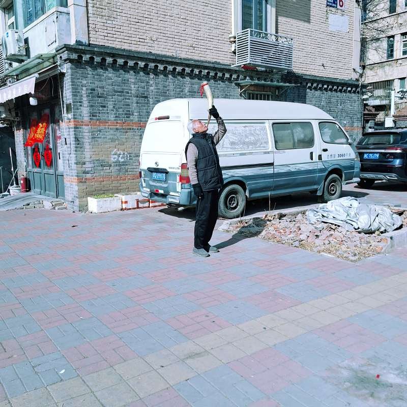 Man standing on a brick-paved sidewalk beside a white and blue van, raising a long curved yellow object toward the sky in an urban street scene.