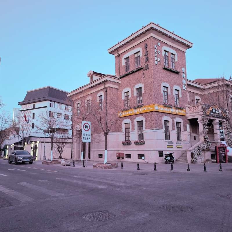 Three-story brick building on a street corner with banners and planters, next to a modern white building, on a quiet urban street.