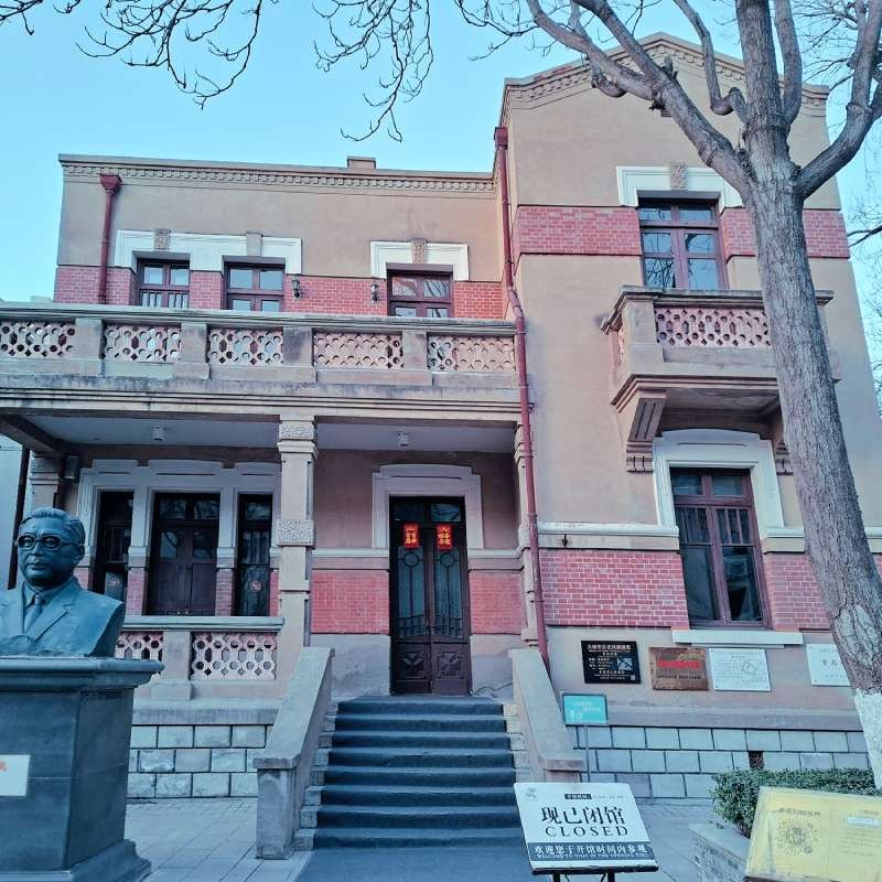 Historic brick-and-stone building with decorative balcony, stairs to a dark door, and a bronze bust statue on a pedestal to the left.