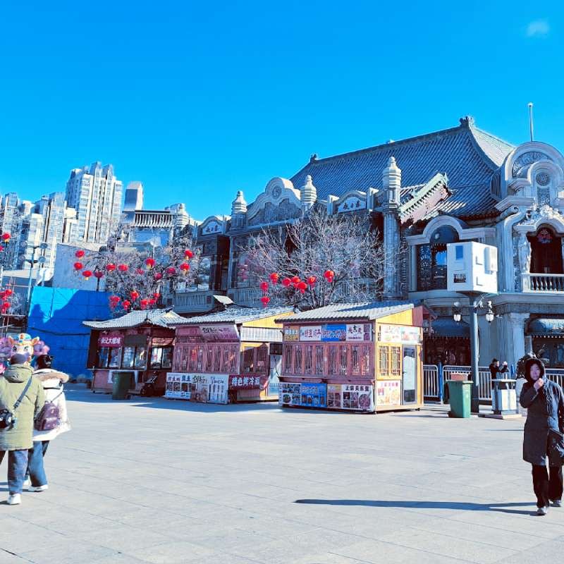 Public square with a traditional Chinese-style building, red lanterns, and market stalls under a clear blue sky.