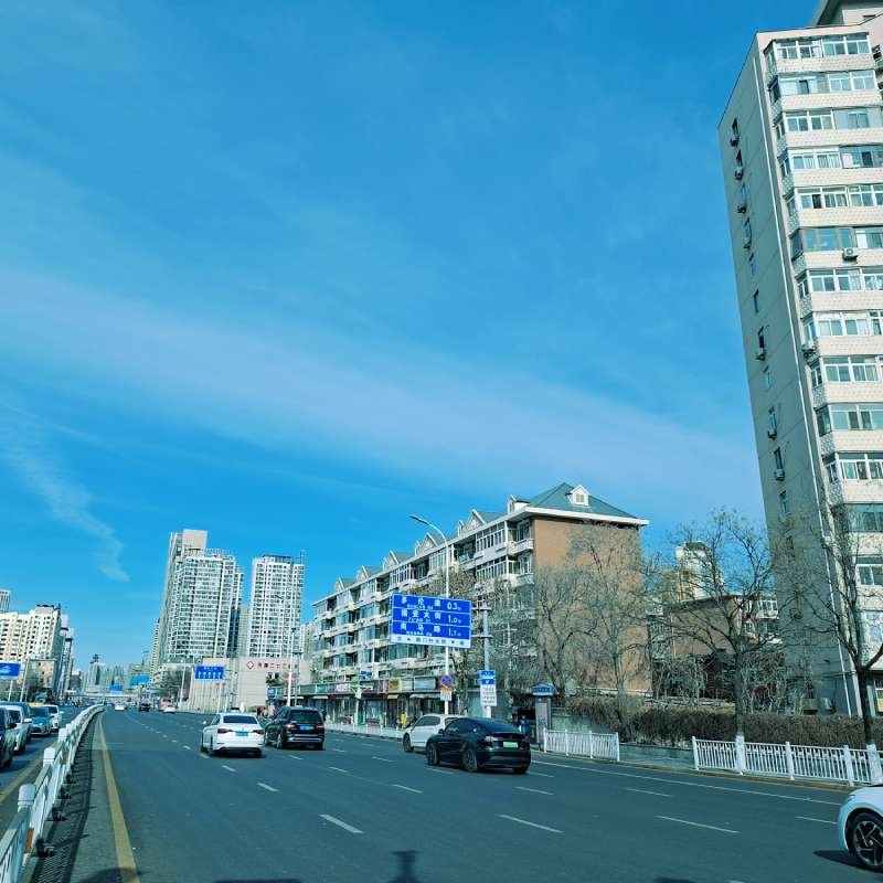 Urban street with tall apartment buildings on the right and a clear blue sky, several cars driving on a multi-lane road.